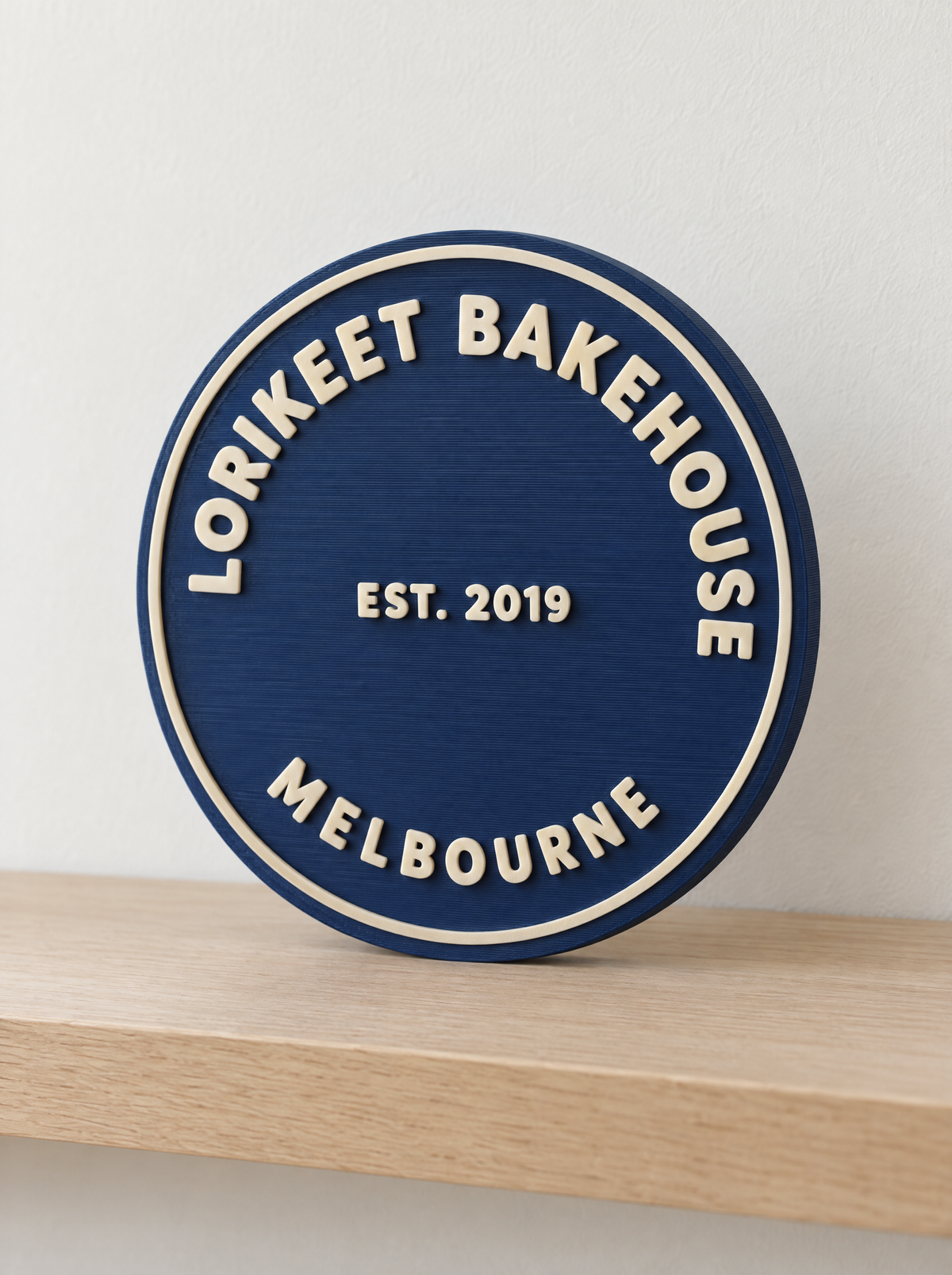 Full Logo Sign in the circle shape: a text-only bakery wordmark 3D printed in cream on a navy disc, standing on a shelf